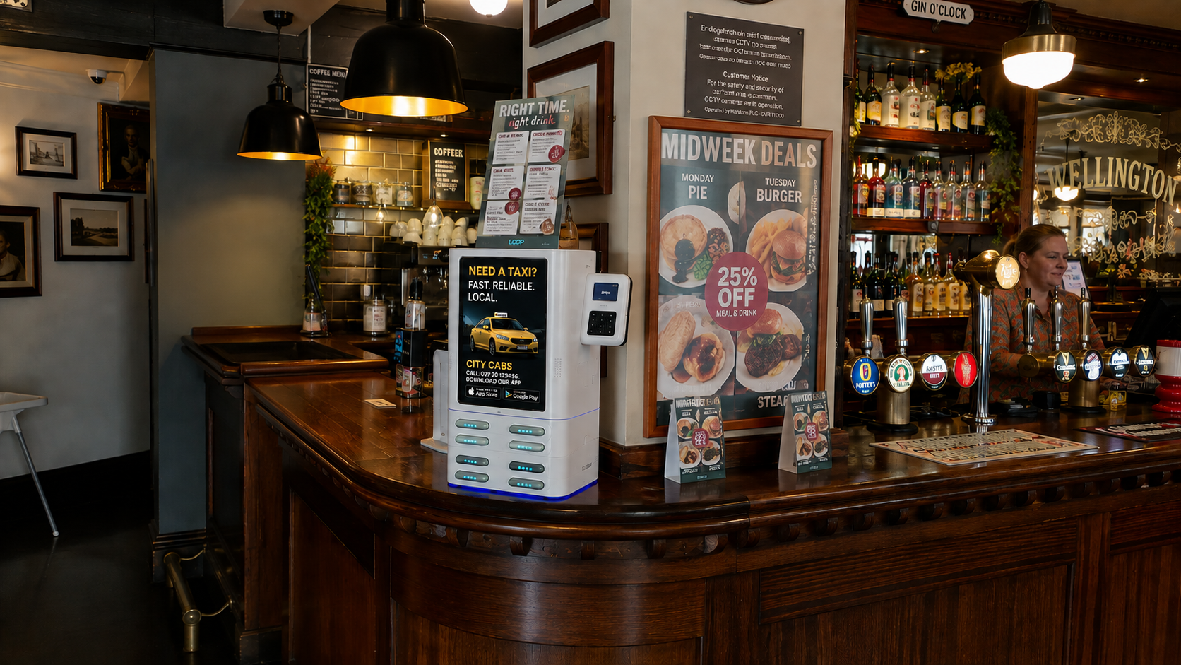 A Loop charging station on the bar of a busy pub, showing a local taxi firm's advert on screen while a member of staff serves behind it.